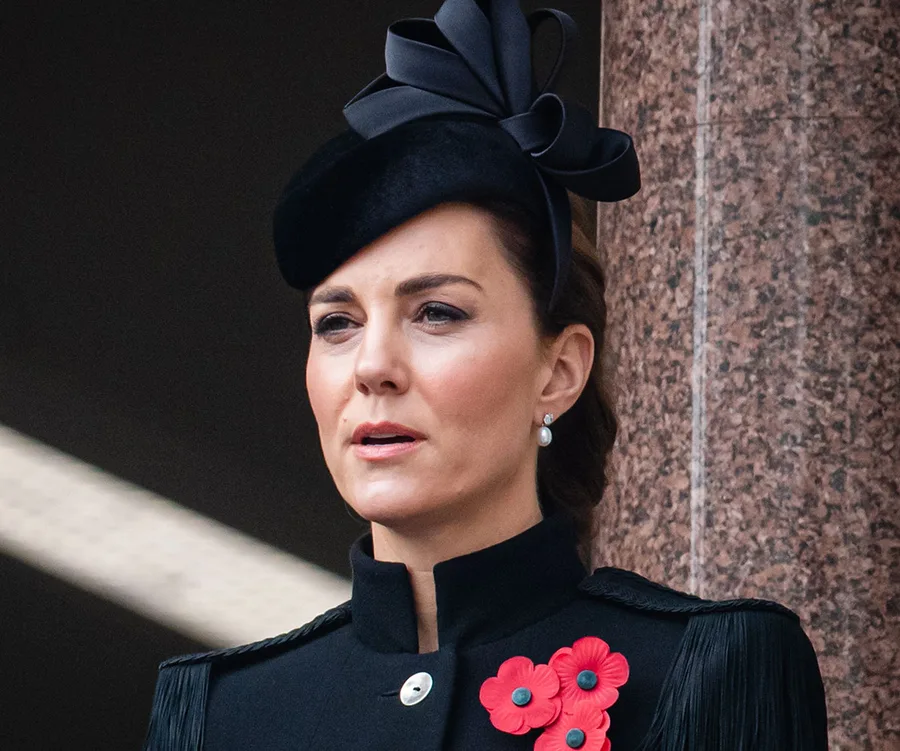 Woman in formal black attire with a decorative black hat and red poppies pinned, standing against a stone wall.
