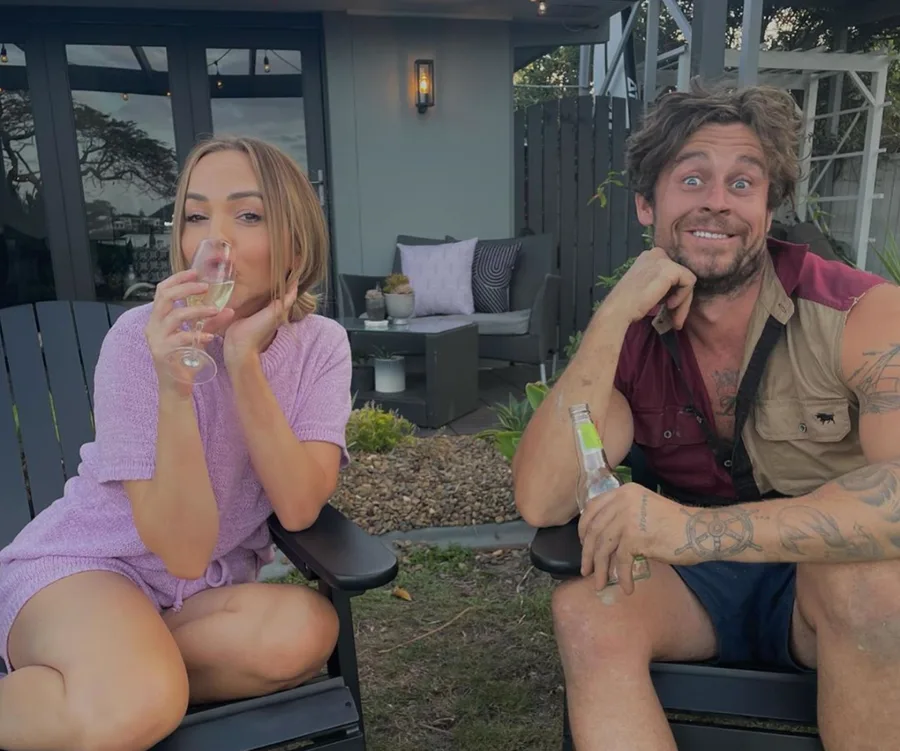 Two people sitting outside, smiling, holding drinks; one sips from a champagne flute, the other from a beer bottle.