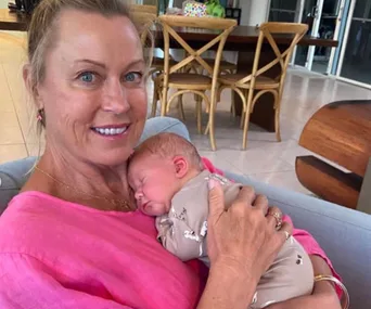 Woman in pink holding a sleeping newborn indoors, with wooden chairs in the background.