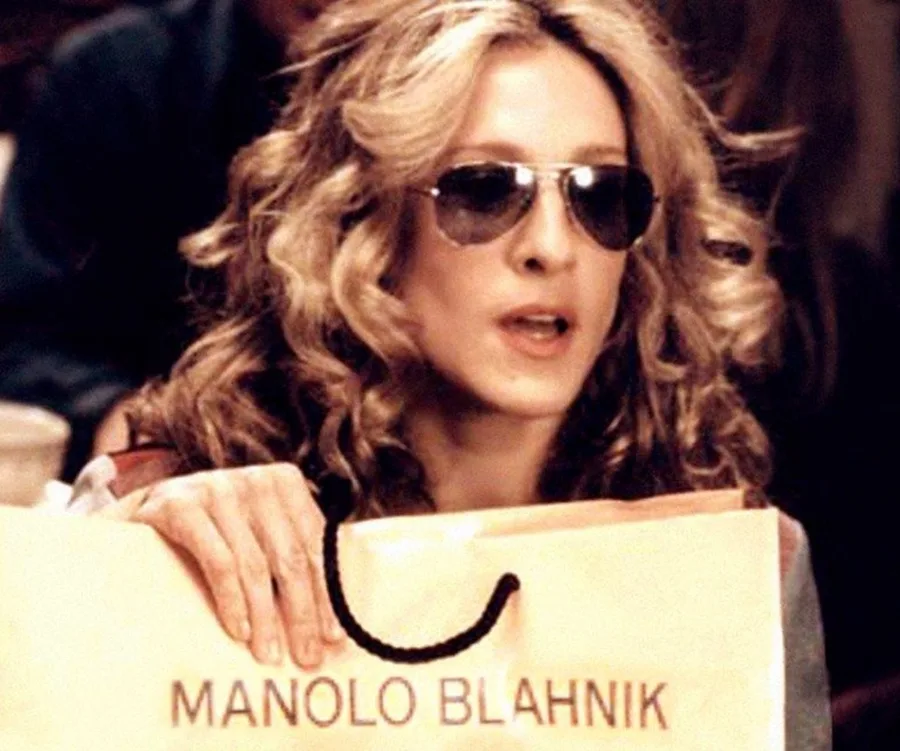 Woman with curly hair, wearing sunglasses, holds a shopping bag labeled "Manolo Blahnik."