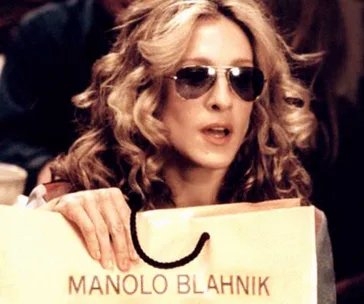 Woman with curly hair, wearing sunglasses, holds a shopping bag labeled "Manolo Blahnik."