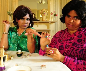 Two women sit at a table with makeup, one in a green dress, the other in pink, holding a mascara wand.