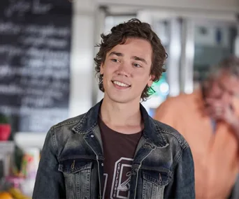 Young man with curly hair in a denim jacket smiling indoors. Blurry background with a person in orange.