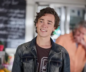 Young man with curly hair in a denim jacket smiling indoors. Blurry background with a person in orange.
