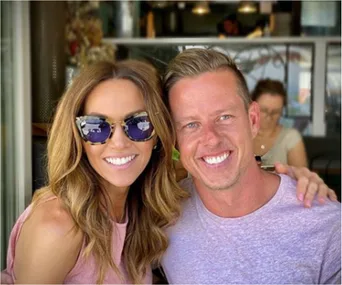 Two people smiling at a cafe, the woman wearing sunglasses and a pink top, the man in a light purple shirt.