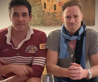 Two men sitting on a couch; one in a maroon shirt, the other in a gray shirt with a blue scarf. A painting is in the background.