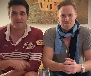 Two men sitting on a couch; one in a maroon shirt, the other in a gray shirt with a blue scarf. A painting is in the background.