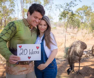 Couple smiling, holding "20 Weeks" sign with red hearts, standing outdoors near emus.