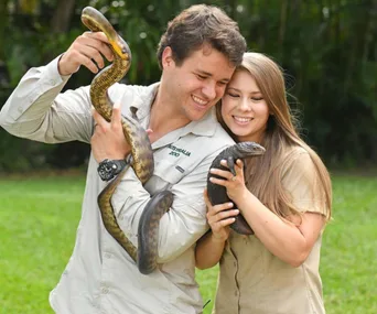 A couple is smiling while holding a snake and a lizard outdoors. They wear khaki shirts, suggesting a wildlife setting.