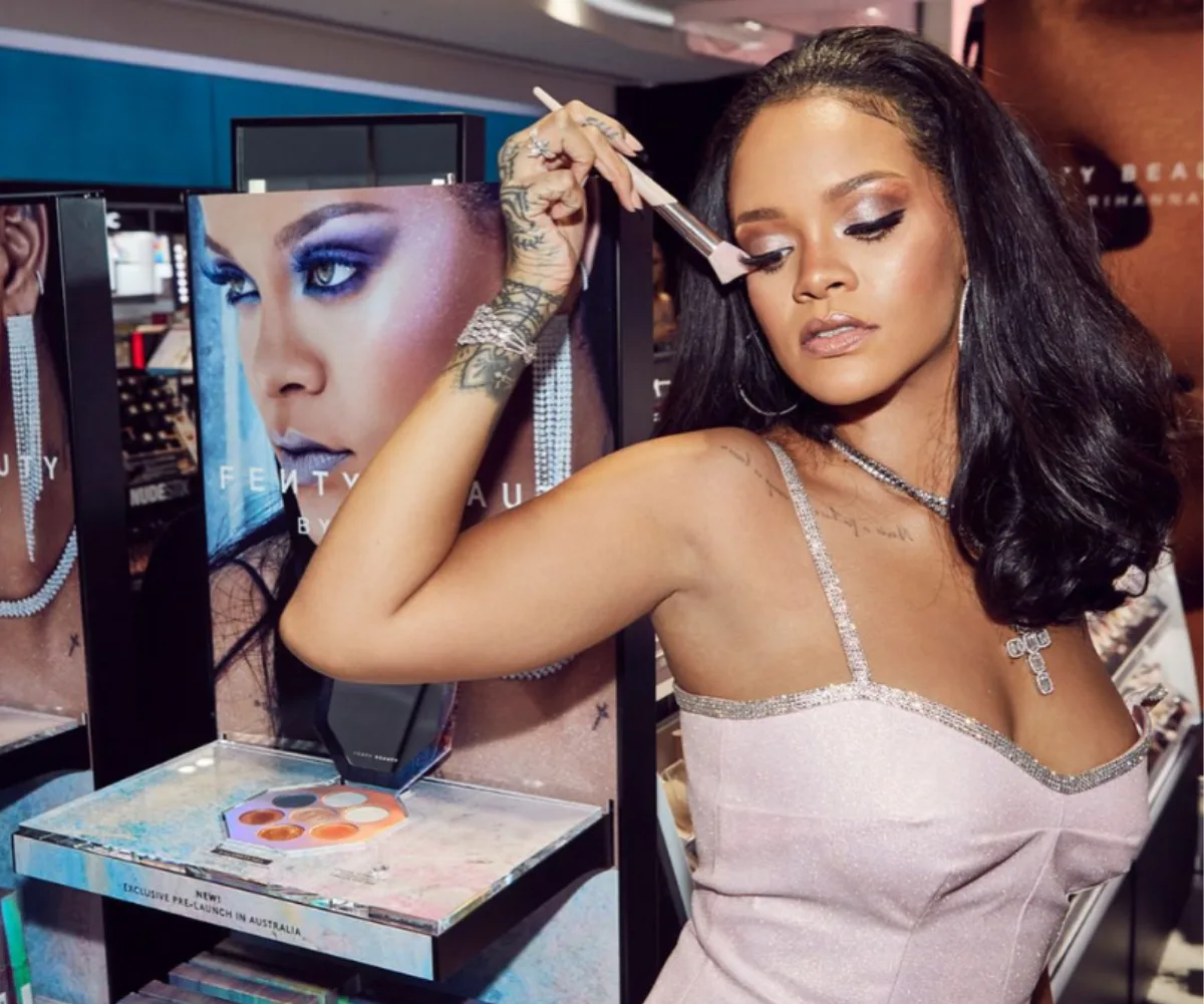 A person uses a makeup brush near a Fenty Beauty display, wearing a shimmering dress and jewelry.