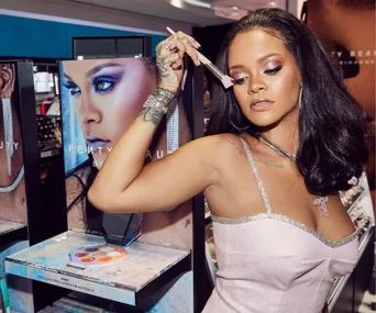 A person uses a makeup brush near a Fenty Beauty display, wearing a shimmering dress and jewelry.