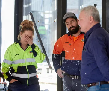 Three people in construction attire look concerned on a site; one woman holds her head, and two men are engaged in conversation.