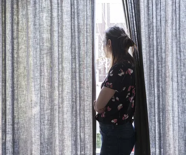 A woman standing by a window, looking outside through curtains, wearing a floral shirt and jeans.