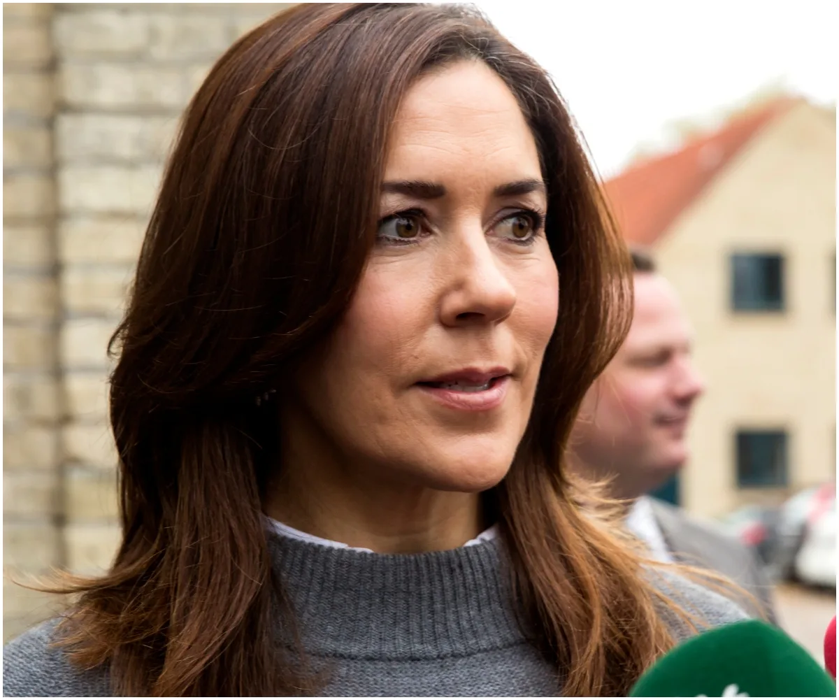 Woman with long brown hair, wearing a gray sweater, speaking at an outdoor event with microphones in the foreground.