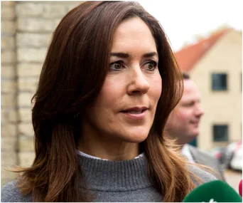 Woman with long brown hair, wearing a gray sweater, speaking at an outdoor event with microphones in the foreground.