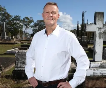 A man in a white shirt stands confidently in a cemetery, surrounded by tombstones and a blue sky.
