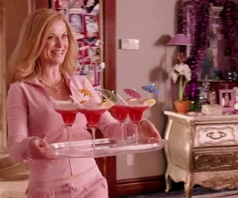 A woman in a pink tracksuit smiles while holding a tray with pink cocktails and umbrellas indoors.