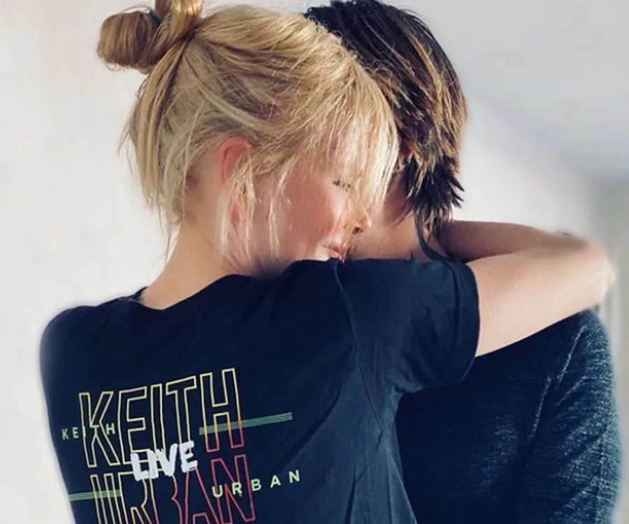 A woman in a "Keith Urban Live" shirt embraces a person with brown hair, in a cozy setting.