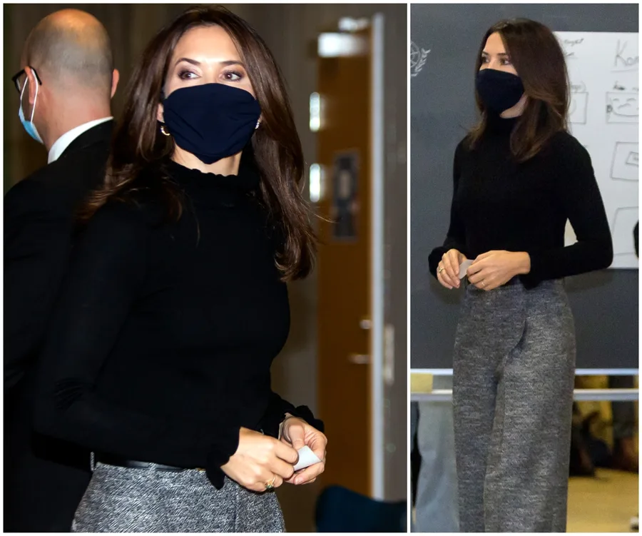A woman in a black top and gray pants wearing a black mask, standing indoors with another masked person in the background.
