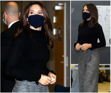 A woman in a black top and gray pants wearing a black mask, standing indoors with another masked person in the background.