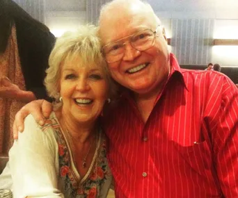 A smiling elderly couple embracing, the man in a red shirt and the woman in a floral blouse.