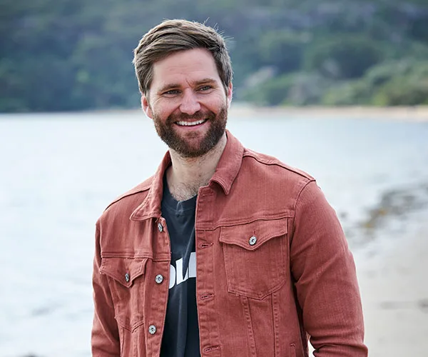 A man with a beard smiles by a shoreline, wearing a rust jacket and dark shirt, in a relaxed outdoor setting.