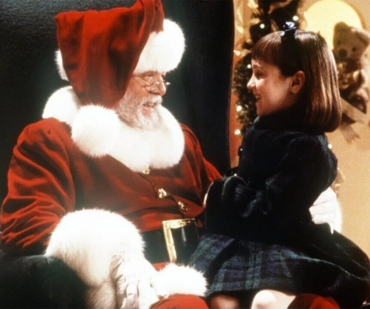 Young girl in a plaid dress sits on Santa's lap, smiling during a festive holiday scene.