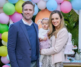 Family standing together at a festive event with colorful balloons and a cake in the background.