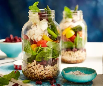 Salad jars with rice, spinach, tomatoes, asparagus, and chicken, next to a bowl of salt on a blue surface.