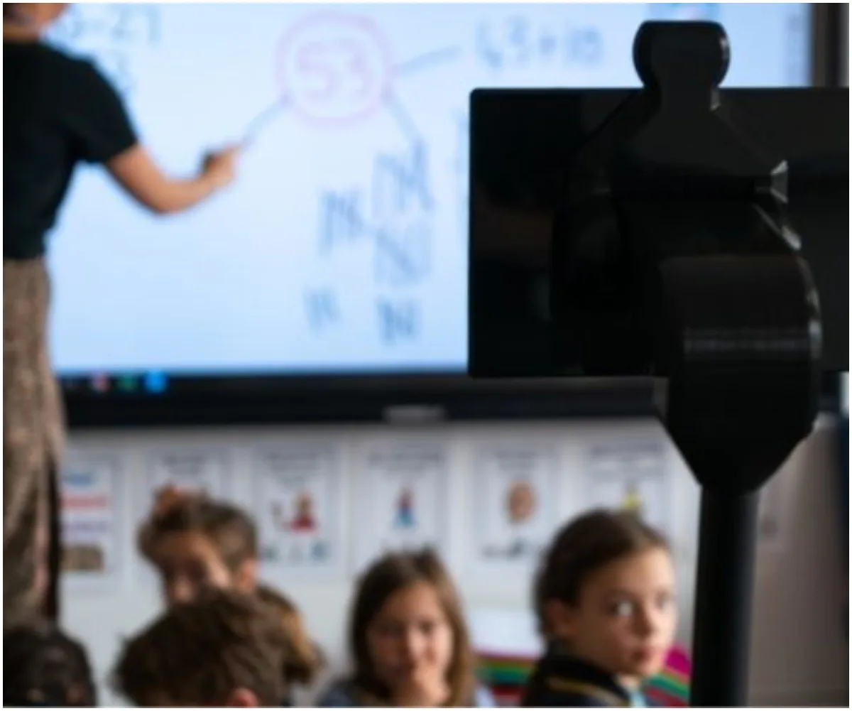 A student writes on a whiteboard in a classroom while a telepresence robot focuses on seated children.