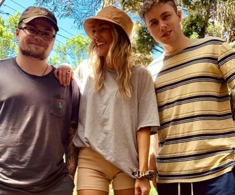 Three people outdoors; one woman between two men, all smiling. Bright day with greenery in the background.