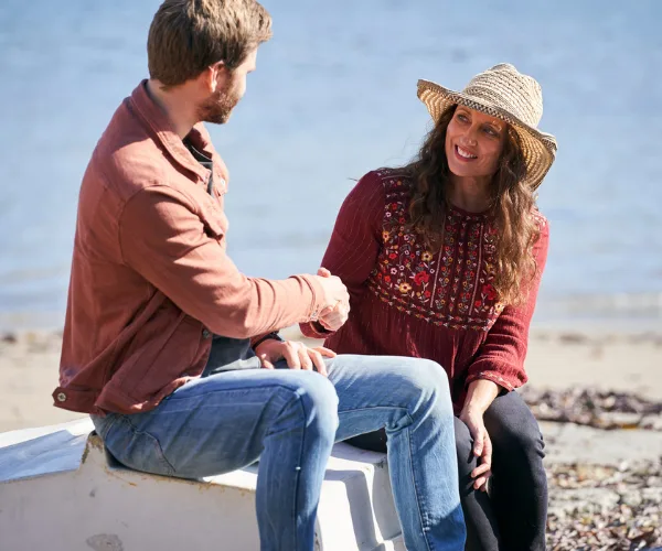 Two people sitting on a boat near a beach, smiling and shaking hands. Woman wearing a hat, both in casual attire.