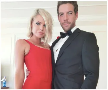 Couple in formal attire: woman in red dress, man in black tuxedo, posing indoors.