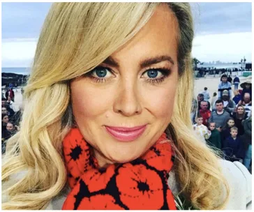 Blonde woman with red and black scarf, smiling at the camera, with a beach and crowd in the background.