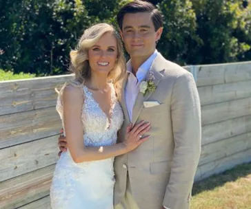 Wedding couple in outdoor setting, bride in lace gown, groom in beige suit with boutonniere, smiling and embracing.