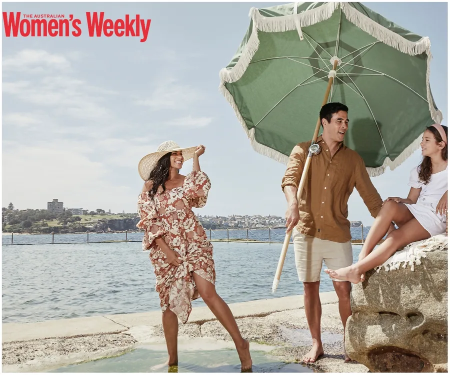 Family enjoying a sunny day by the water; woman in floral dress, man holding umbrella, girl sitting on rock.