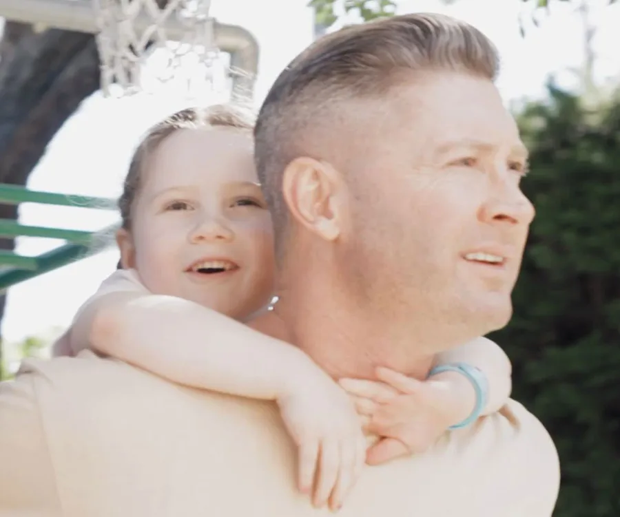 Father and daughter outdoors, child on father's back, smiling and enjoying the moment under a clear sky.