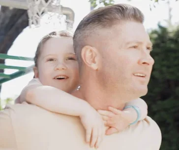 Father and daughter outdoors, child on father's back, smiling and enjoying the moment under a clear sky.