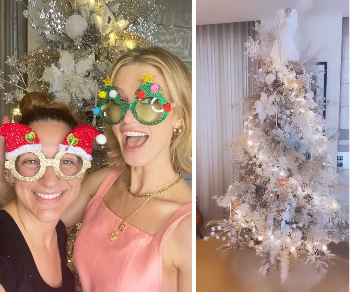 Two women wearing festive glasses pose by a glittering Christmas tree with lights and an angel topper.