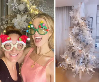 Two women wearing festive glasses pose by a glittering Christmas tree with lights and an angel topper.