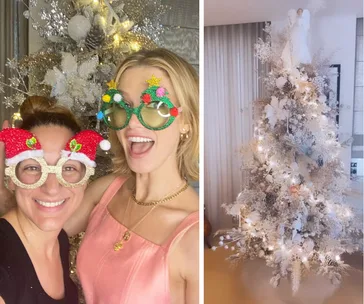 Two women wearing festive glasses pose by a glittering Christmas tree with lights and an angel topper.