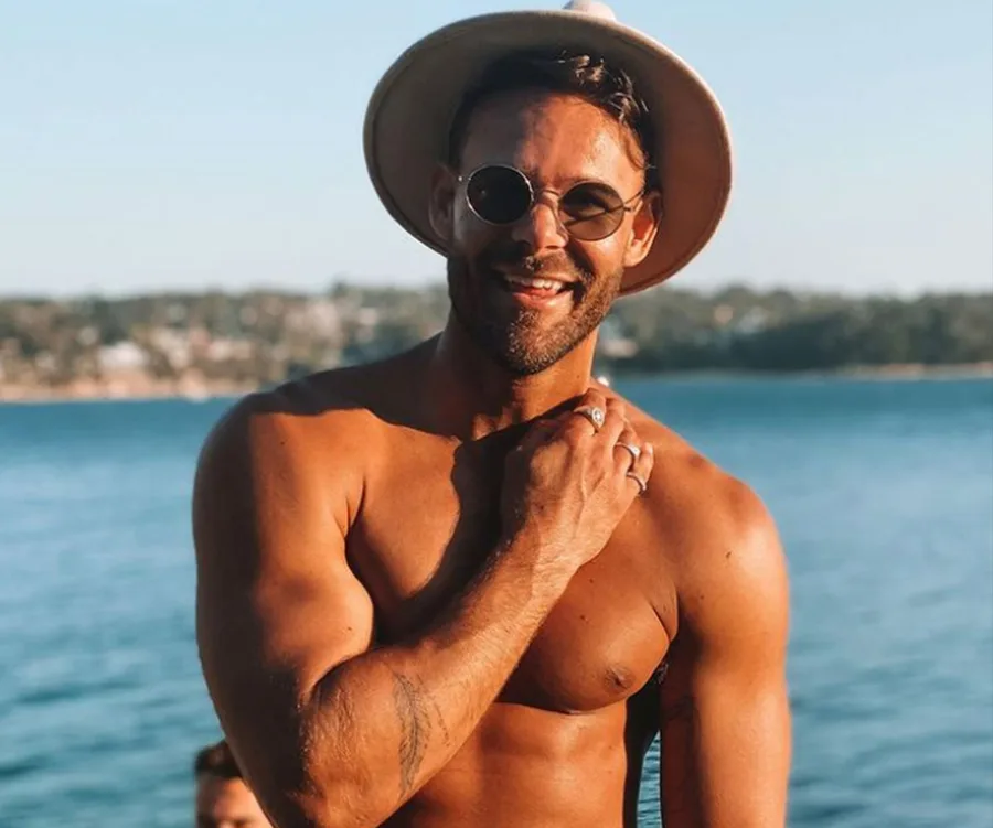 A man wearing sunglasses and a hat smiles by the water, showing off his muscular physique on a sunny day.