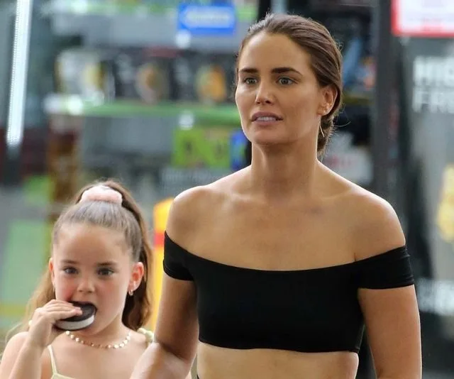 Woman in black off-shoulder top with a child eating a snack, standing indoors.