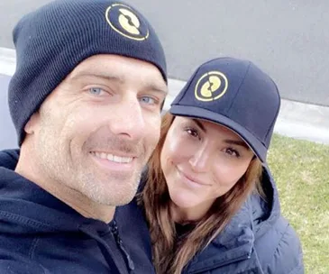 Two people wearing black hats with matching logos, smiling outdoors.