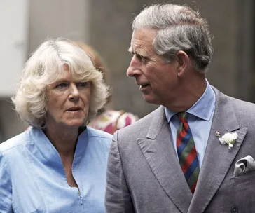 A man in a gray suit with a striped tie stands beside a woman in a blue blouse, both appear to be outdoors.