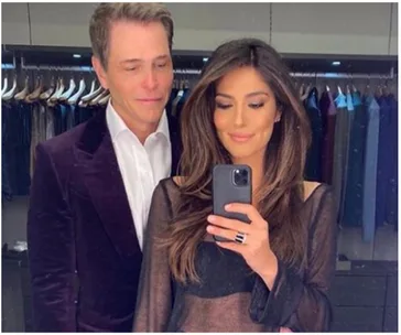 A couple taking a mirror selfie in a closet with clothing hanging in the background. The woman holds the phone.