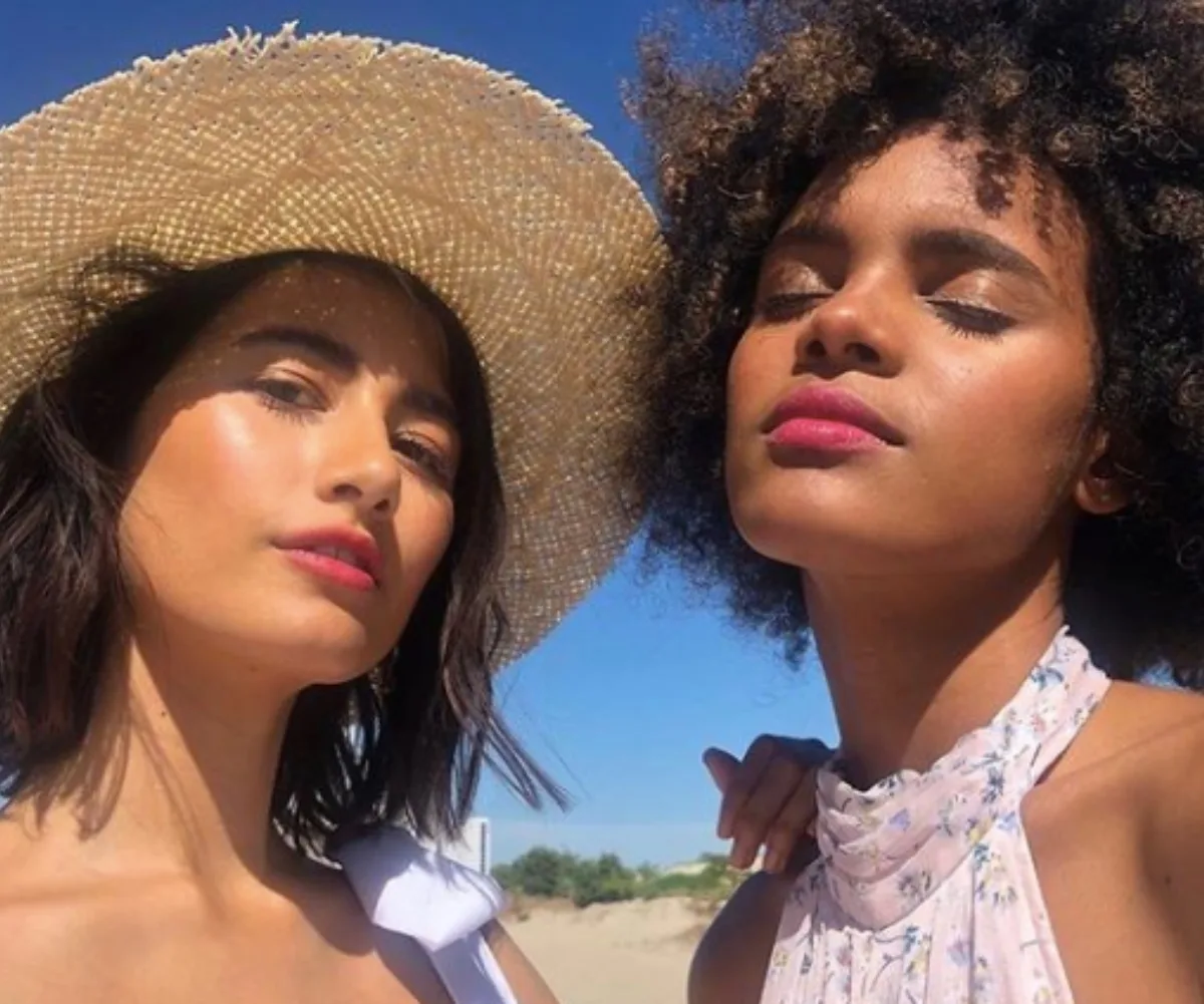 Two women outdoors, one with a straw hat, both with natural makeup, enjoying the sunlight.