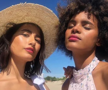 Two women outdoors, one with a straw hat, both with natural makeup, enjoying the sunlight.