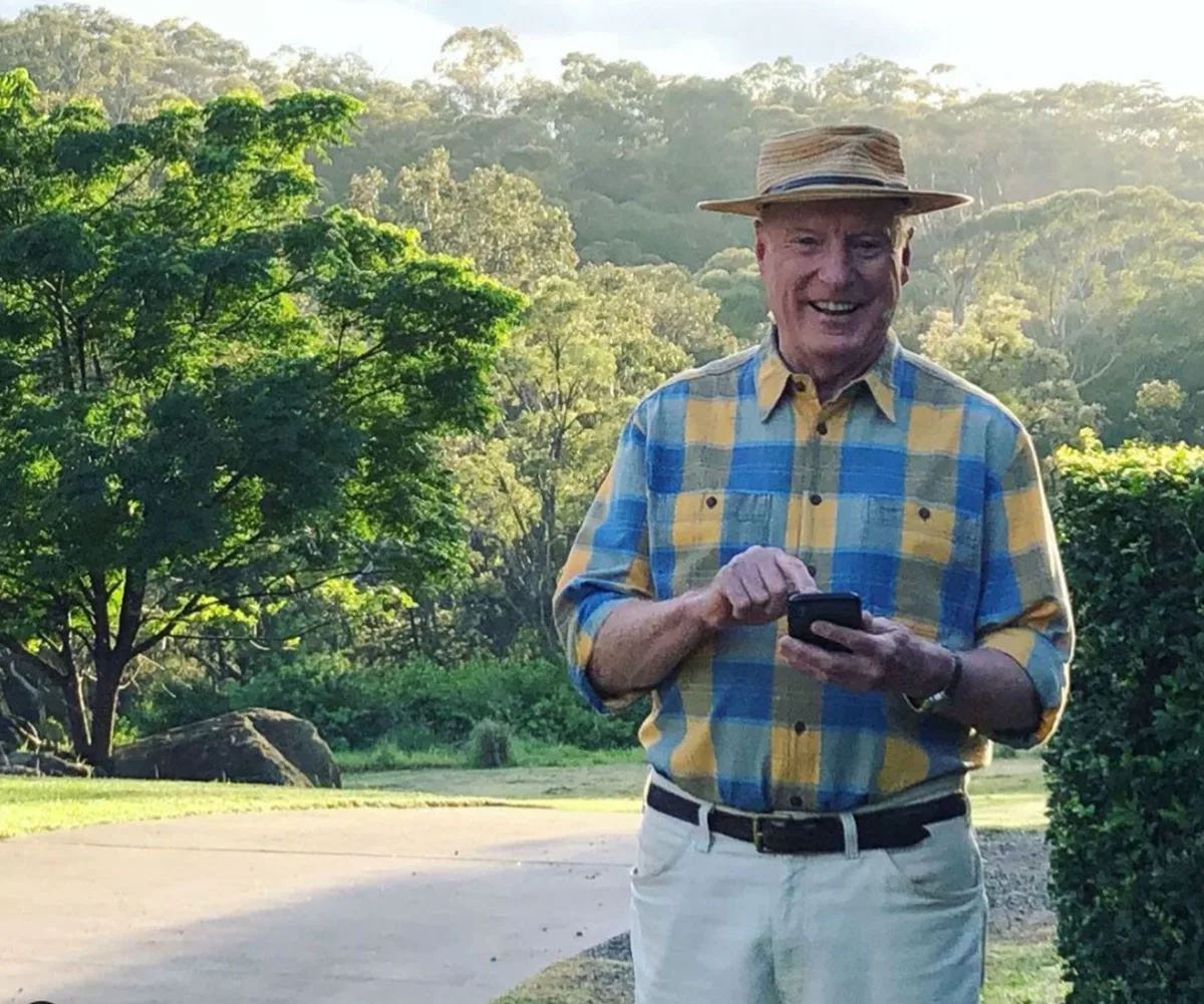 Man in a yellow and blue checkered shirt and straw hat, smiling and using a smartphone in a sunny, wooded area.