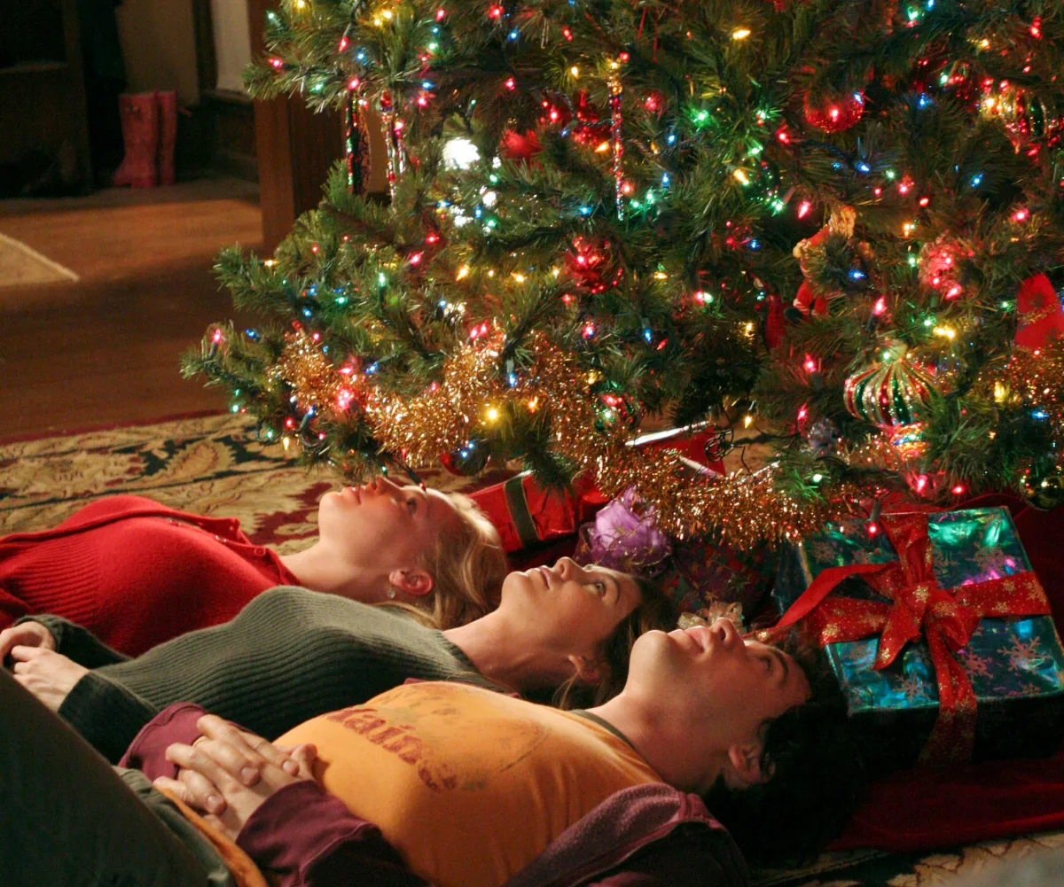 Three people lie on the floor looking up at a decorated Christmas tree with colorful lights and wrapped presents underneath.